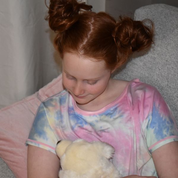 Child with Red Hair in buns sitting on a couch looking at her toy. She is wearing a tie dyed sensory friendly nightdress