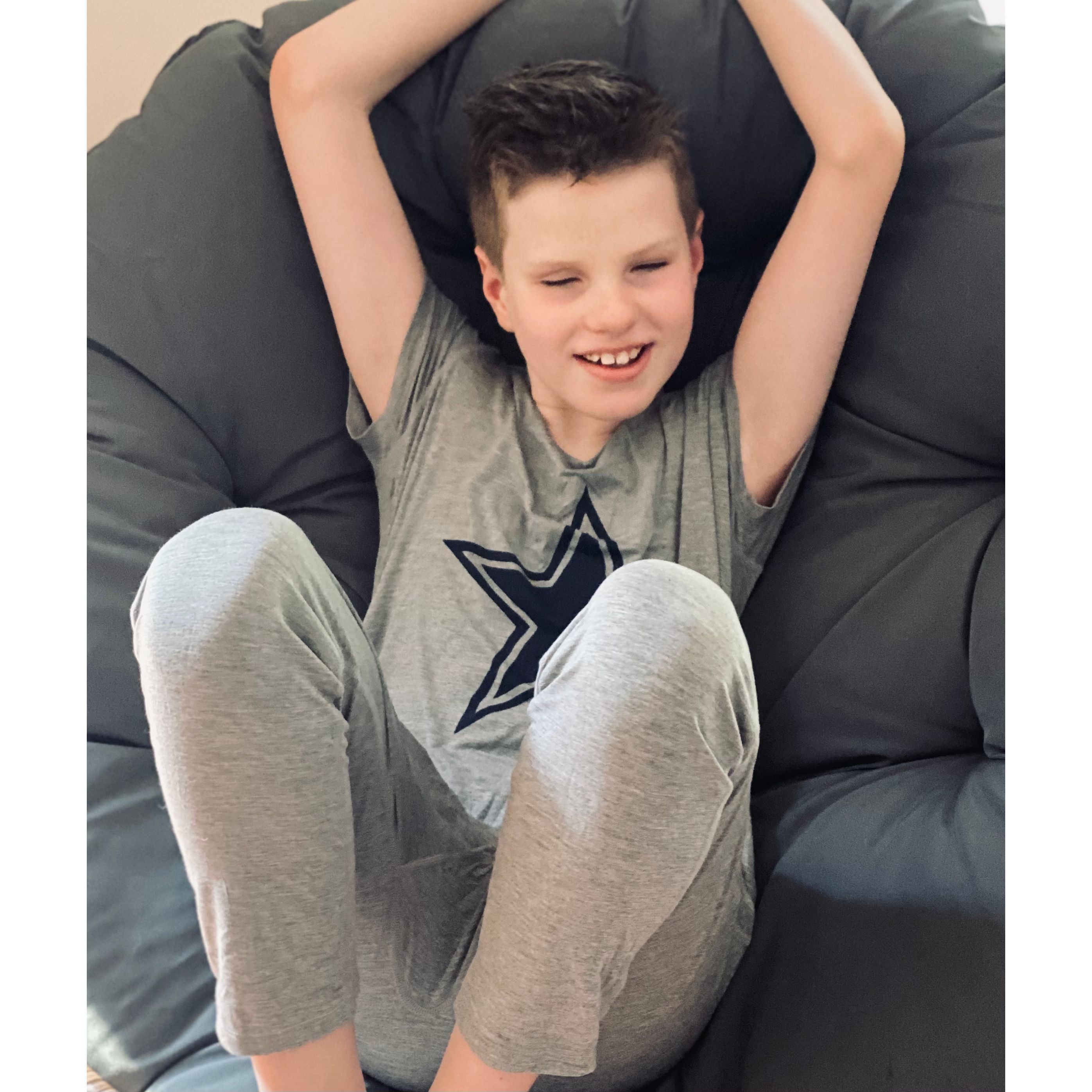 A sitting and smiling boy wearing the Grey Marle Jumpsuit Pajamas for kids and teens from Comfort on the Spectrum.