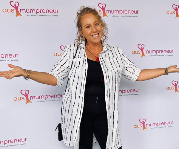 Picture of Kelly wearing black jeans, top & white striped shirt. She is standing in front of a white board that has Ausmumpreneur stamped on it