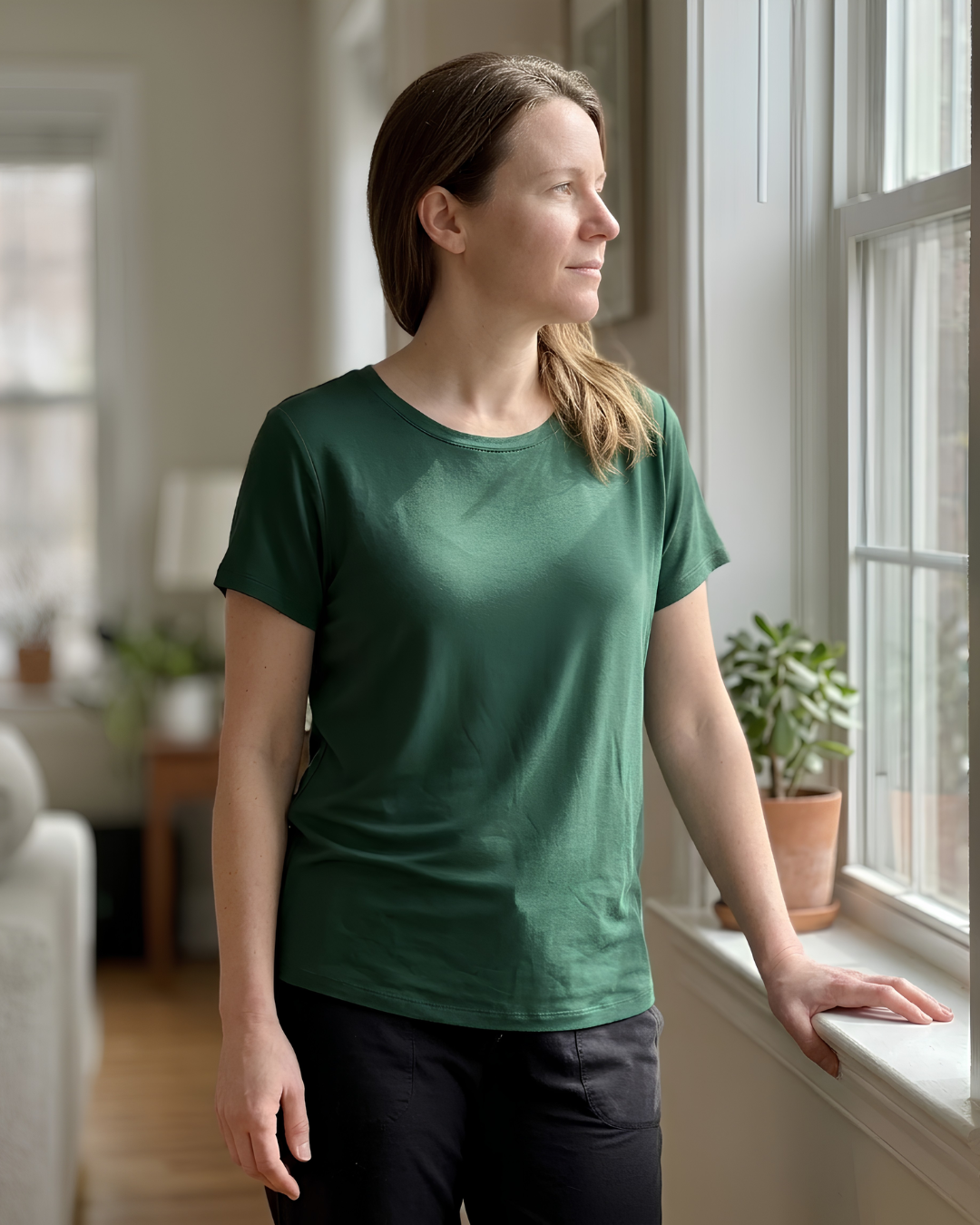 A woman wearing a dark green short-sleeve crewneck t-shirt and black pants, standing by a window in a bright, modern room.