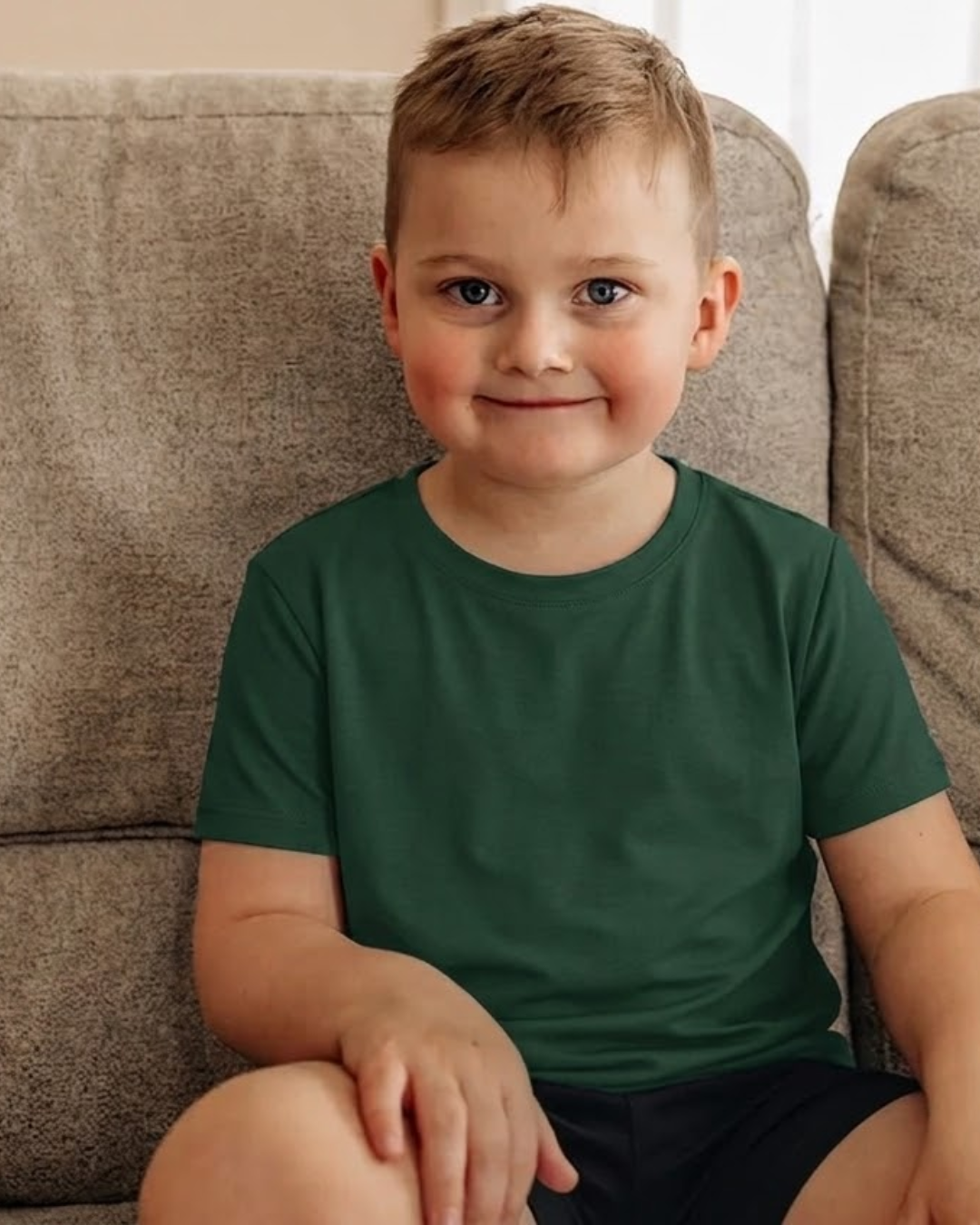 A young boy with short blond hair sits on a textured gray couch, wearing a solid dark green crew-neck t-shirt and dark shorts.