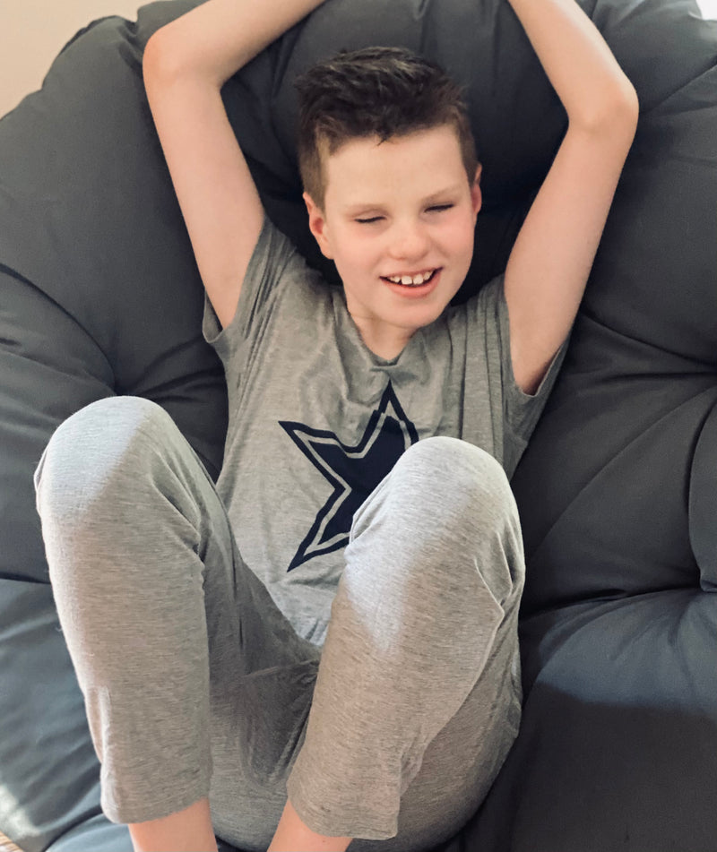 A sitting and smiling boy wearing the Grey Marle Jumpsuit Pajamas for kids and teens from Comfort on the Spectrum.